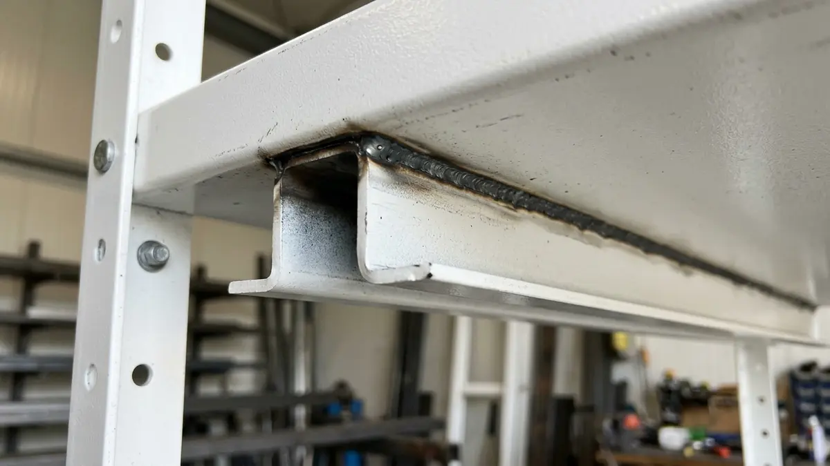 Visão detalhada em close da parte inferior de uma bandeja de aço branca de gôndola comercial, destacando um perfil metálico em formato de canal ômega soldado longitudinalmente para reforço de carga.
